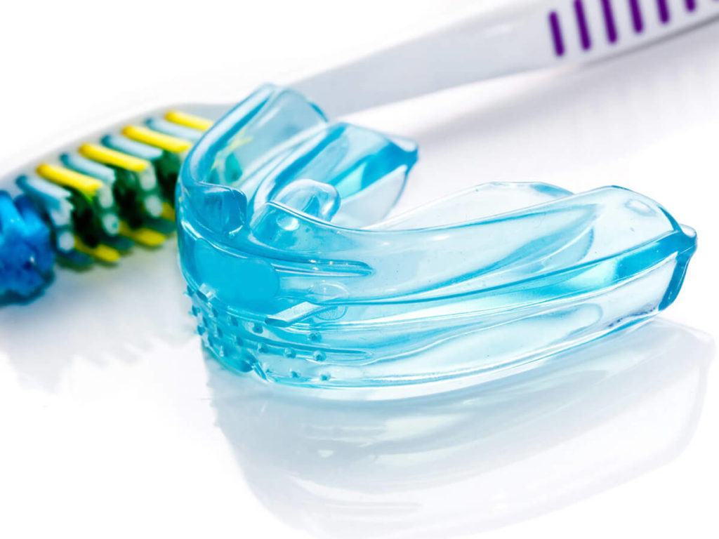 Close-up image of a clear blue dental mouthguard on a white background, with a multicolored toothbrush blurred in the background, illustrating dental care and protection equipment.