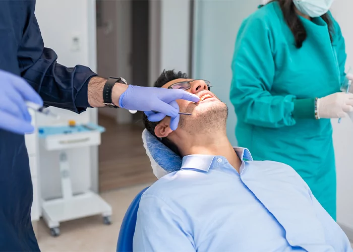 A patient reclining in a dental chair receives a local anesthesia injection in his mouth from a dentist wearing blue gloves, with a dental assistant in green scrubs preparing equipment in the background, depicting a calm and professional dental treatment environment.