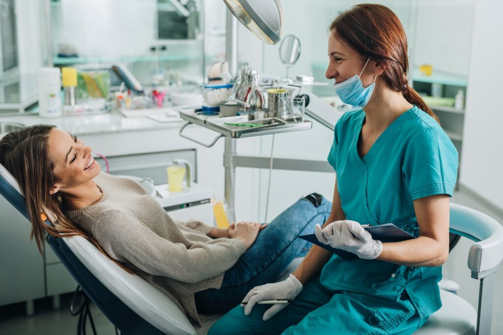 A cheerful female patient reclines in a dental chair, smiling at her female dentist who is wearing a teal scrubs and a surgical mask, holding a clipboard in a modern, well-equipped dental clinic, suggesting a positive and communicative dental visit.