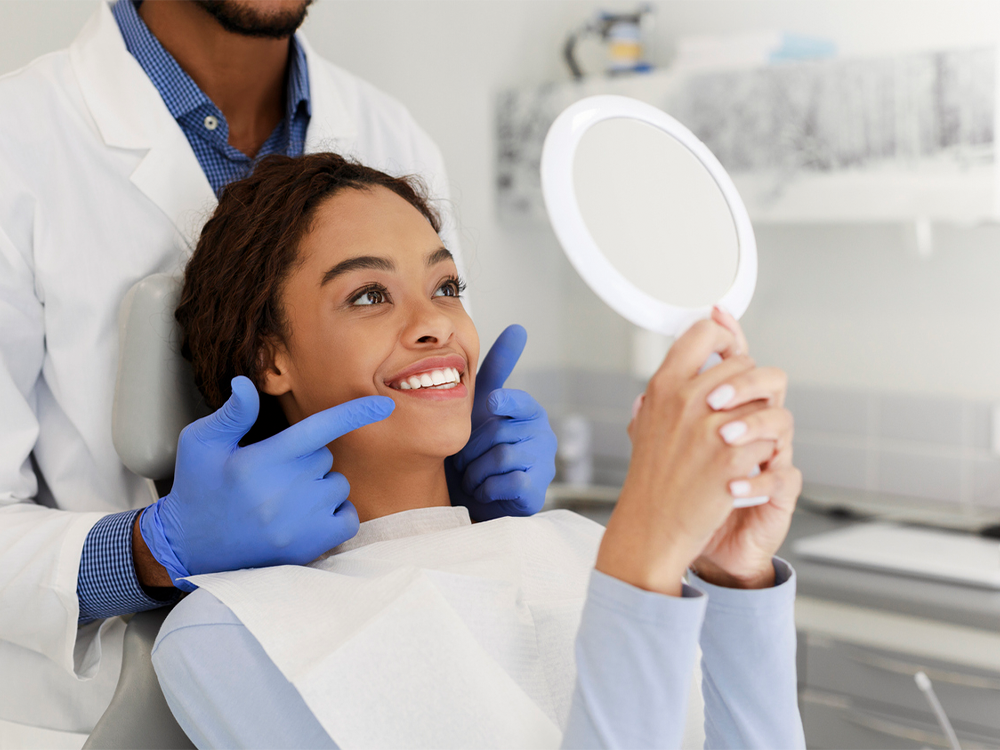A happy young woman in a dental chair looking at her smile in a hand-held mirror, while a male dentist in a white coat and blue gloves points to her teeth, indicating a successful dental procedure in a bright and modern dental clinic.