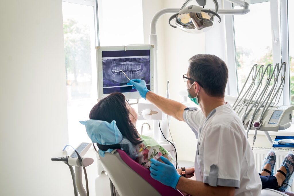 A female patient in a dental chair looks at her dental X-ray on a monitor while a male dentist, wearing a mask and pointing with a pen, explains the details. The clinic is well-lit with modern dental equipment in the background, emphasizing a professional dental consultation.