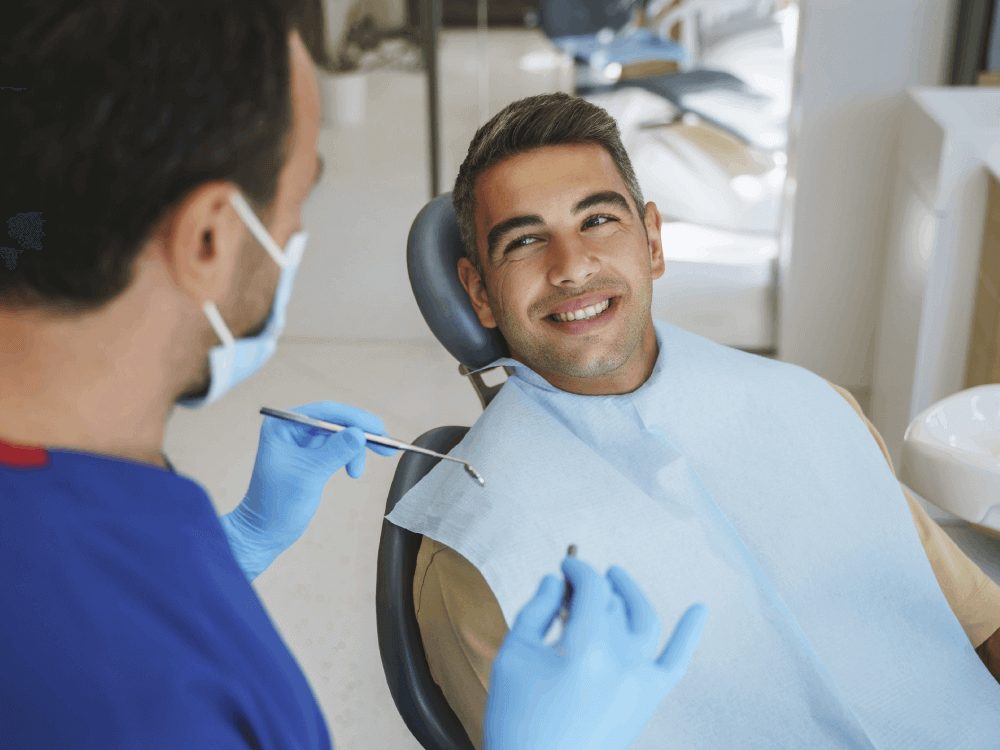 A cheerful young man smiling in a dental chair during a consultation with his male dentist, who is wearing blue gloves and a mask, in a modern dental clinic setting, portraying a positive and professional dental care experience.