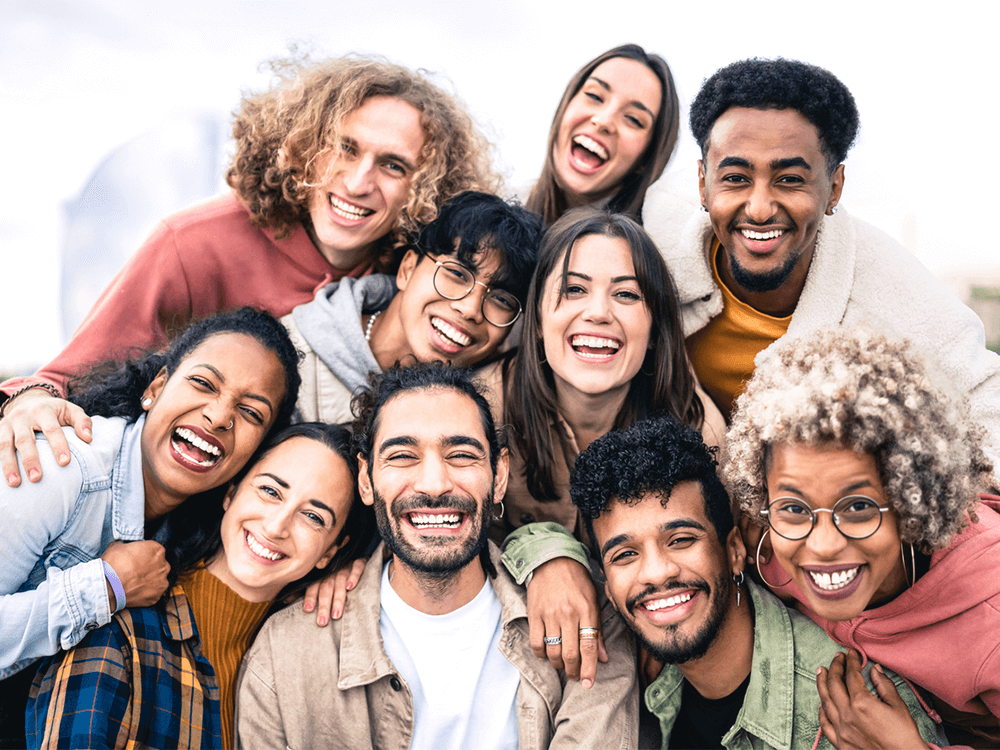 Joyful group portrait of eight diverse friends outdoors, smiling broadly. The image captures a mix of men and women from various ethnic backgrounds, expressing happiness and togetherness, suggesting a positive social gathering