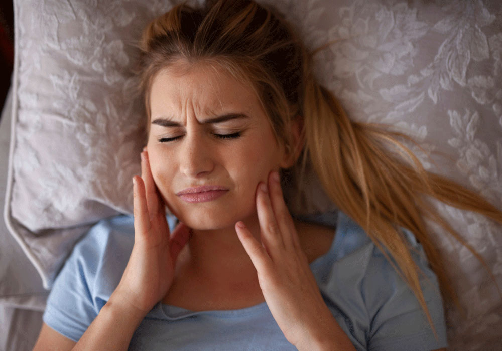A young woman lying in bed, clutching her cheeks and grimacing in pain, suggesting symptoms of TMJ or toothache, with a background of ornate pillows enhancing the nighttime discomfort scenario.